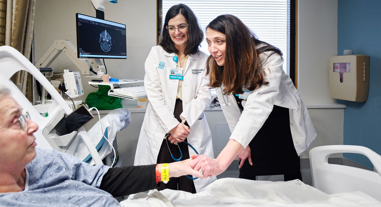 An older patient lies in a hospital bed while two medical professionals in white coats stand beside the bed. One doctor gently holds the patient's hand. Medical equipment and a monitor displaying a brain scan are visible in the background.