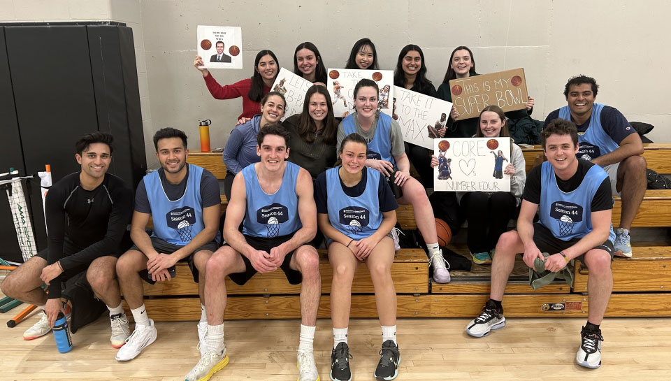 On a low set of bleachers, the CORE Lab team, in light blue jerseys, sit surrounded by friends. Their friends hold up signs cheering them on.