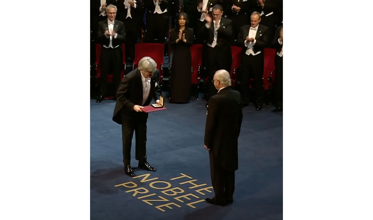 A bowing man presents the Nobel Prize to a second man as the audience gives a standing ovation.