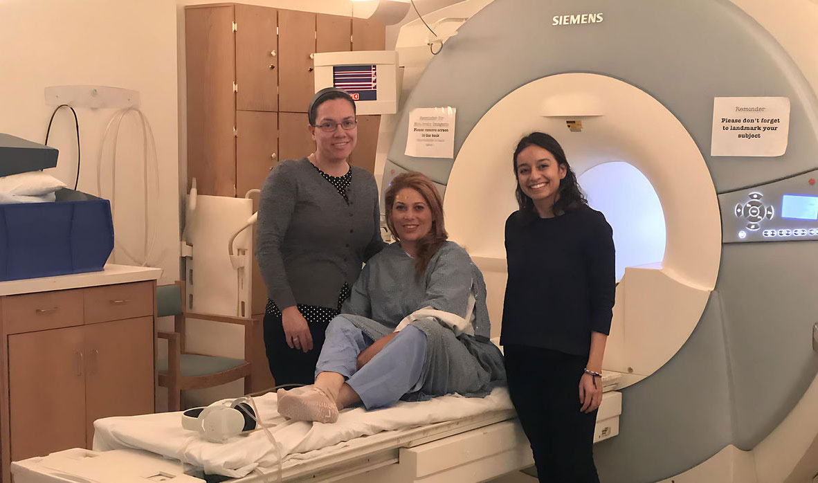Three women posing beside an MRI machine, with one woman sitting on the MRI bed.