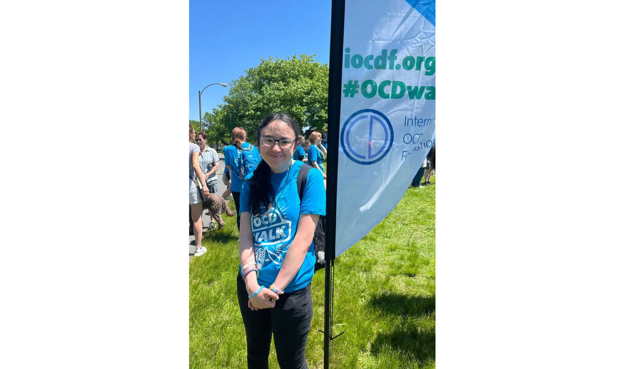 A young girl in a blue shirt that says OCD Walk, standing beside a banner for the OCD Walk.