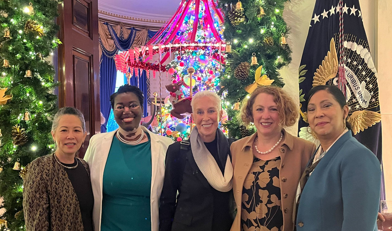 Five women posing in fromt of a doorway that's decorated for Christmas, with a presidential flag beside them and a stunning pink merry-go-round made of glass ornaments in the room behind them.