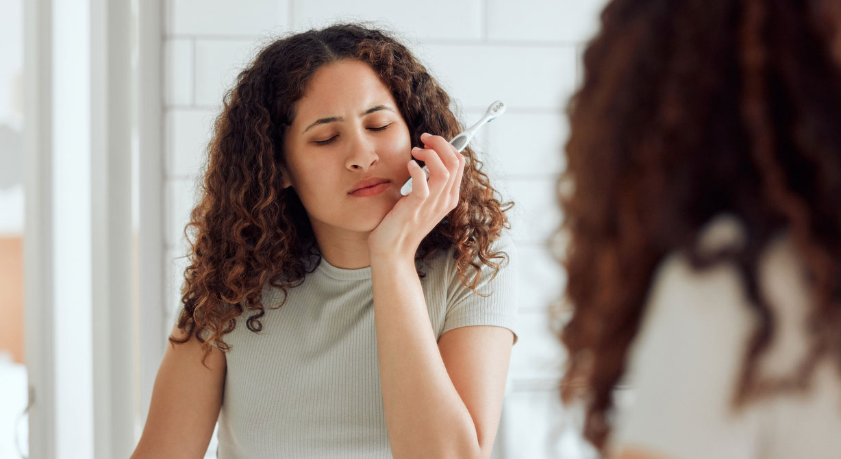 A woman with a toothbrush in her hand holds her jaw like her teeth hurt.