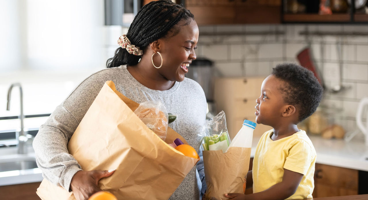 A mom and young son smile at each other as they place paper bags full of healthy groceries on the kitchen counter. 
