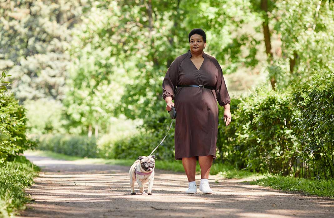 An overweight woman walks her pug in the park.