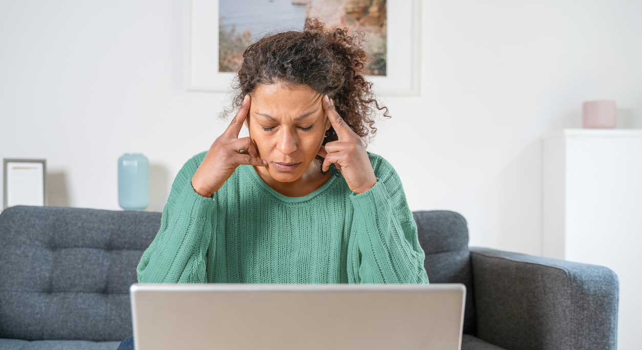 A woman sits on her sofa in front of a laptop, holding her head like she has a headache.