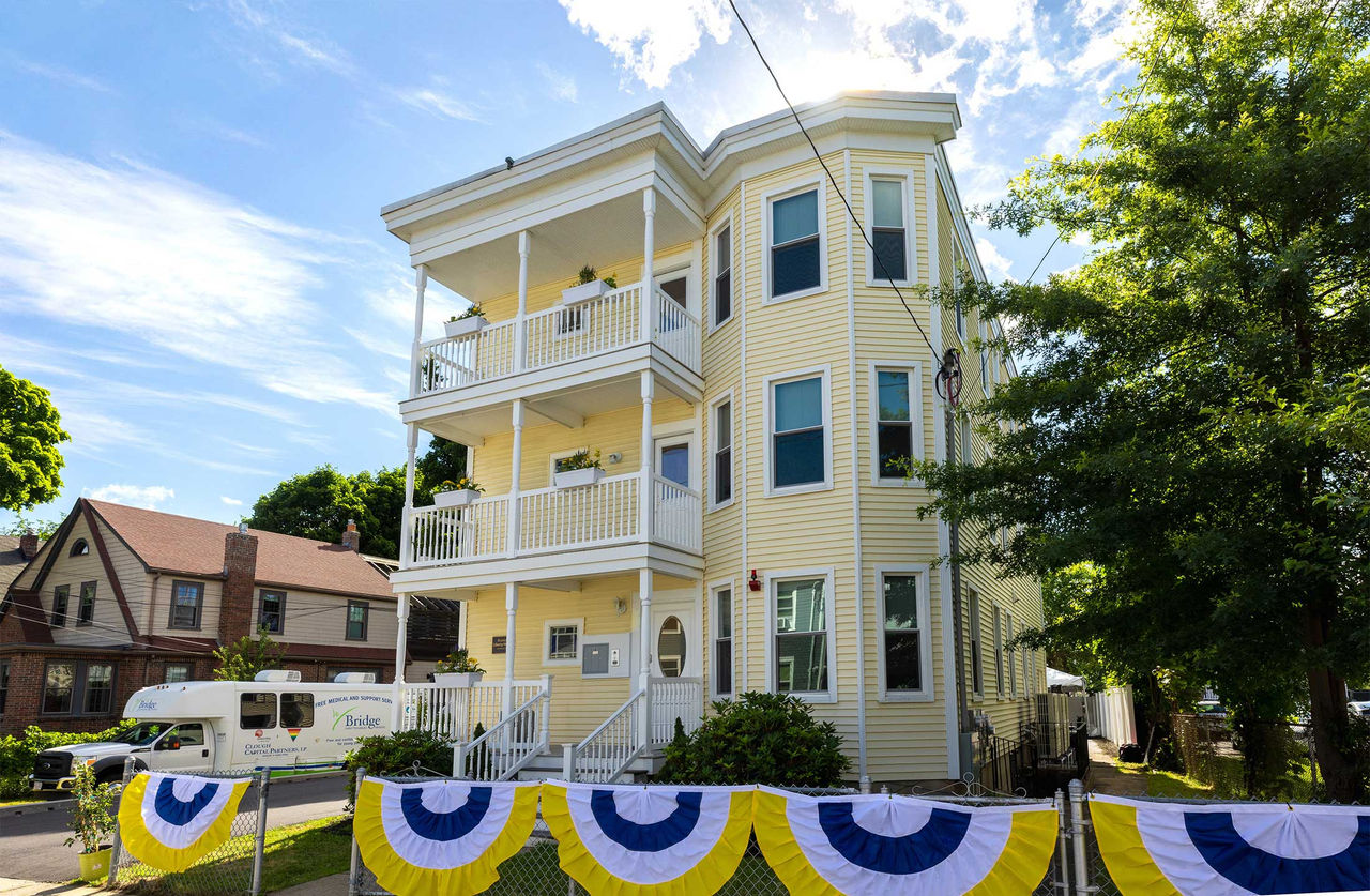 A bright yellow triple-decker house with white trim. Yellow, white, and black bunting hangs on the front fence.