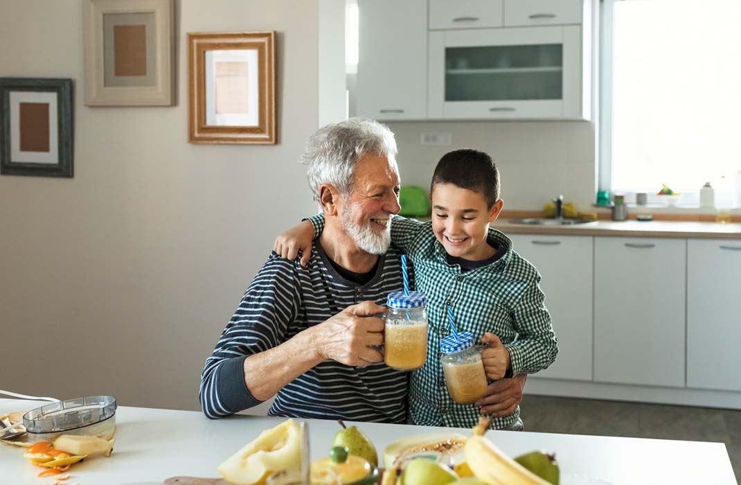 A grandfather with white hair and beard dressed in a black long sleeve striped top smiling and enjoying a healthy drink with his grandson who has black hair and is wearing a long sleeve black gingham button-up shirt with a collar. Both of them are in sitting in a kitchen with cabinets in the back and a kitchen table in the front with  fruits on it.