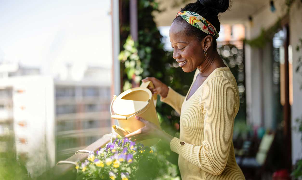 A smiling woman waters plants on a balcony.