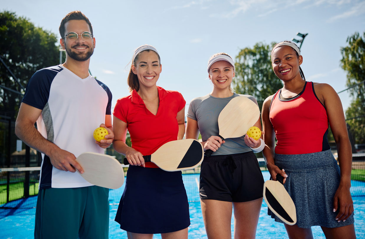A multi-racial group of young adults with pickleball paddles smiling after a pickleball game.