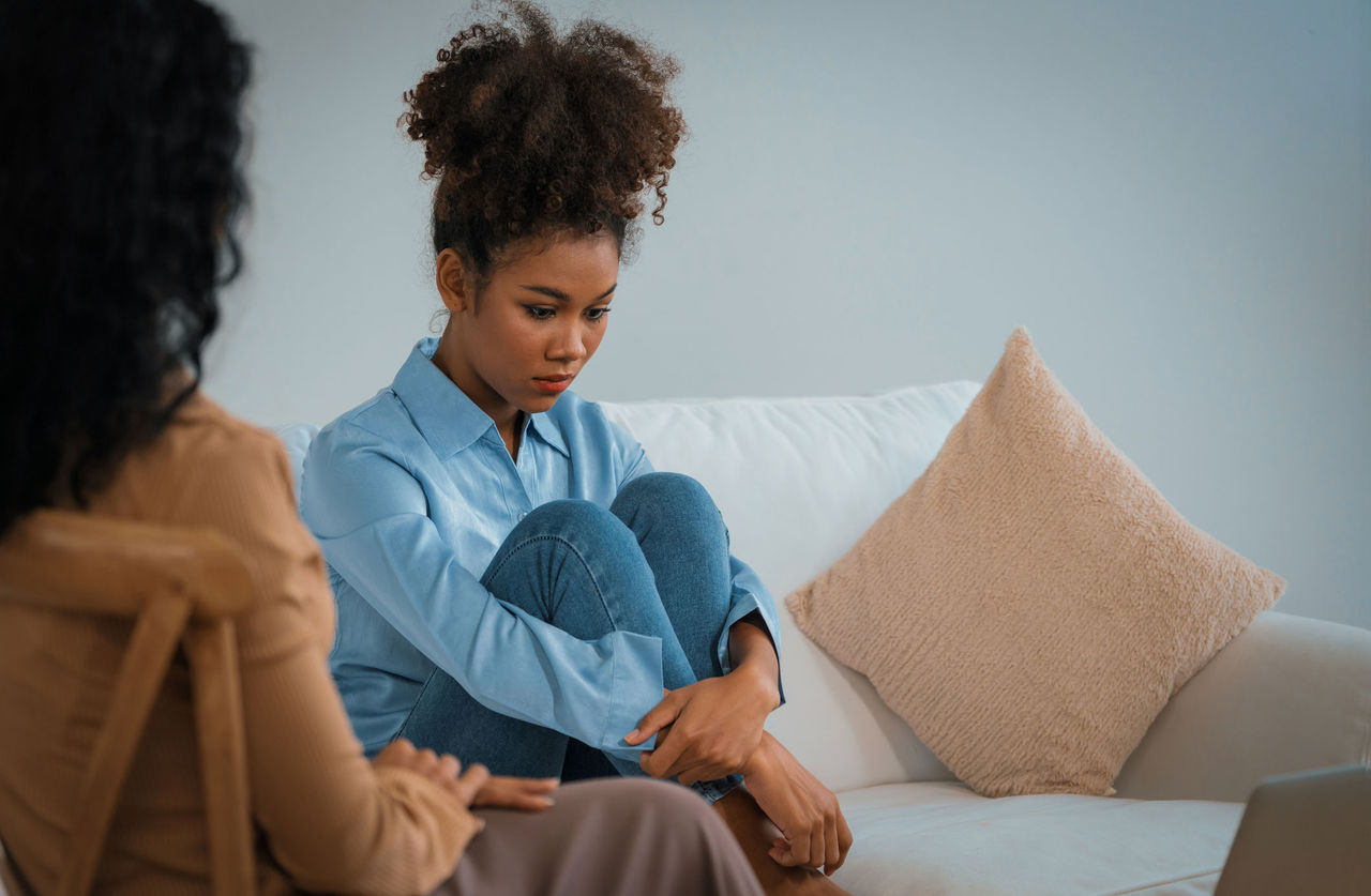 A sad woman hugs her knees on a sofa while her therapist talks to her.