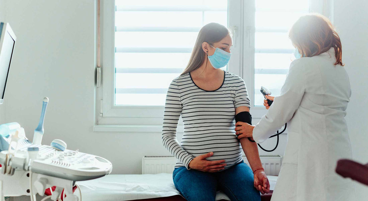 A medical professional in a face mask checks the blood pressure of a pregnant woman in a face mask as she is seated on an examination table