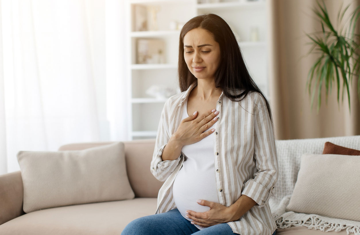A pregnant woman touches her chest in discomfort, sitting on a couch at home.