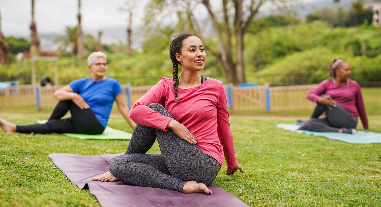 Three women do a yoga stretch in a green park.