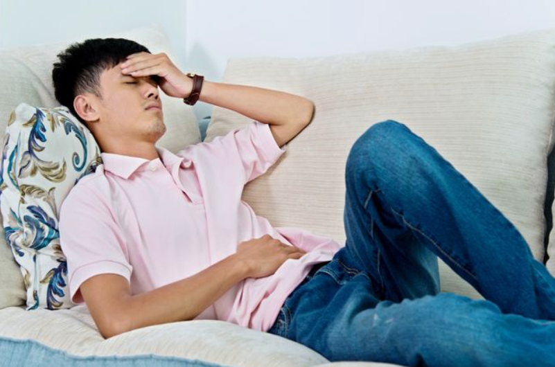 A grimacing man lies on a sofa, holding a hand to his forehead and stomach.