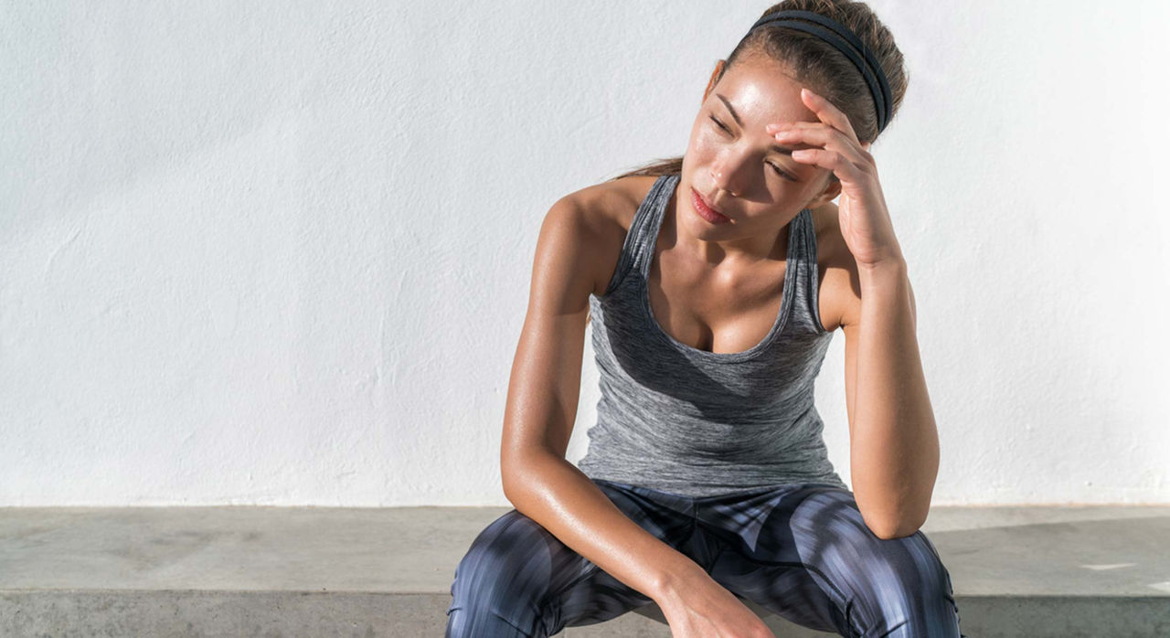 An athlete in workout gear sitting and resting on a concrete bench with their head resting on their left hand after exercising in the heat