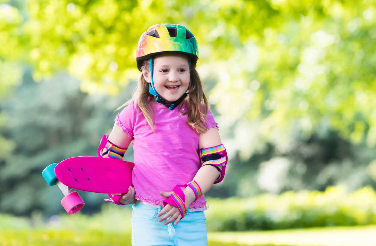 A smiling young girl holds her skateboard in the park, wearing pink and rainbow-colored protective gear.