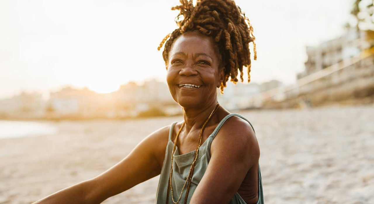 A smiling, confident older woman wears a tank top and sits on the beach at sunset.