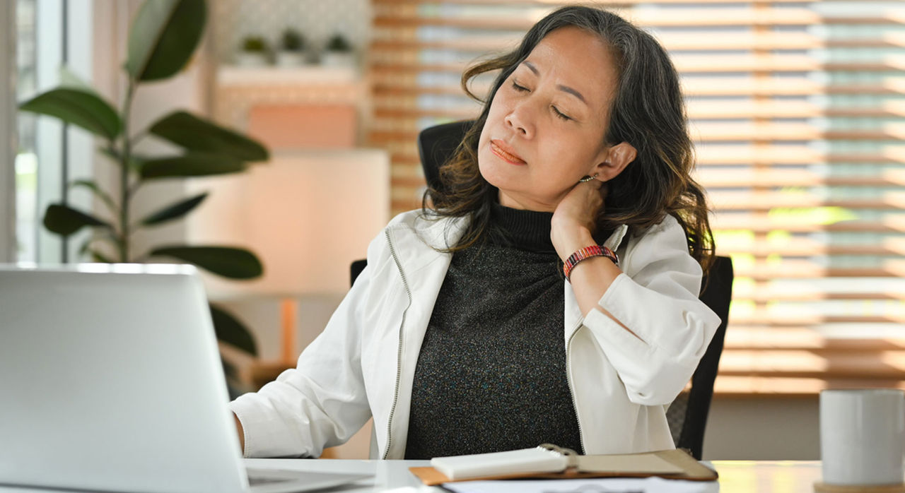 A woman touches her neck in discomfort from tech neck, sitting at a desk with her laptop.