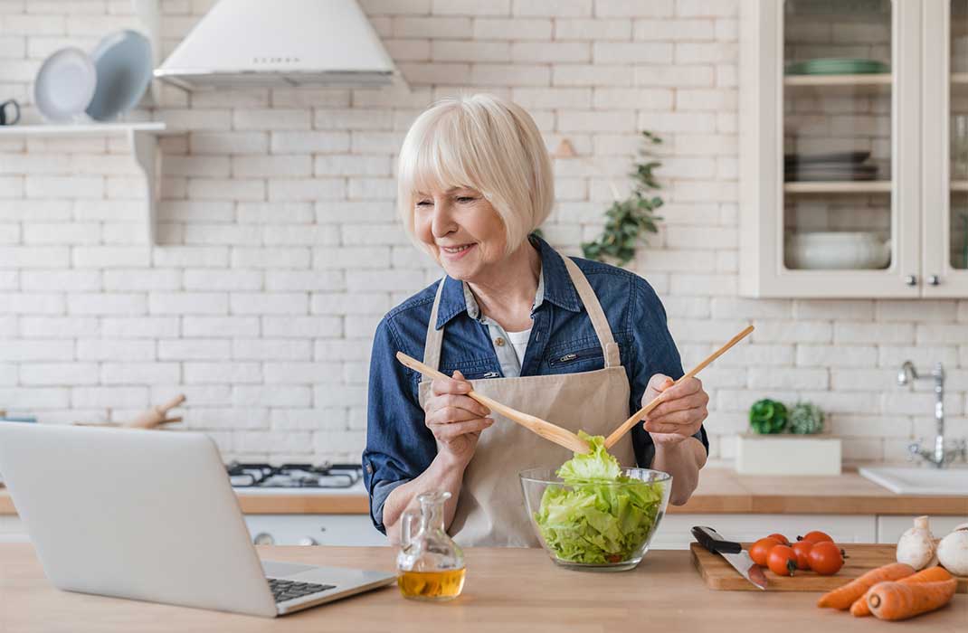 An older woman consults her laptop while making a salad.