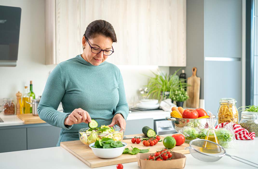 A woman in a longsleeve top wearing glasses with her hair pulled back puts together a salad with lots of ingredients set out on the kitchen counter