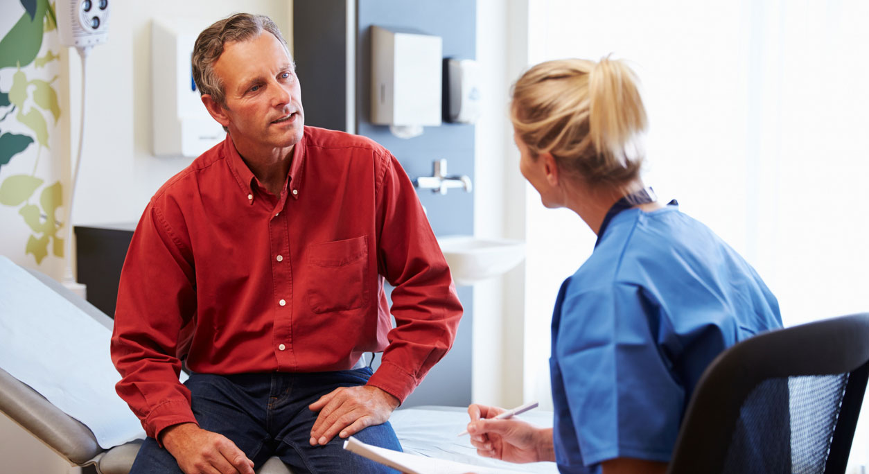 A man sits on an exam table and discusses health screening options with a provider.
