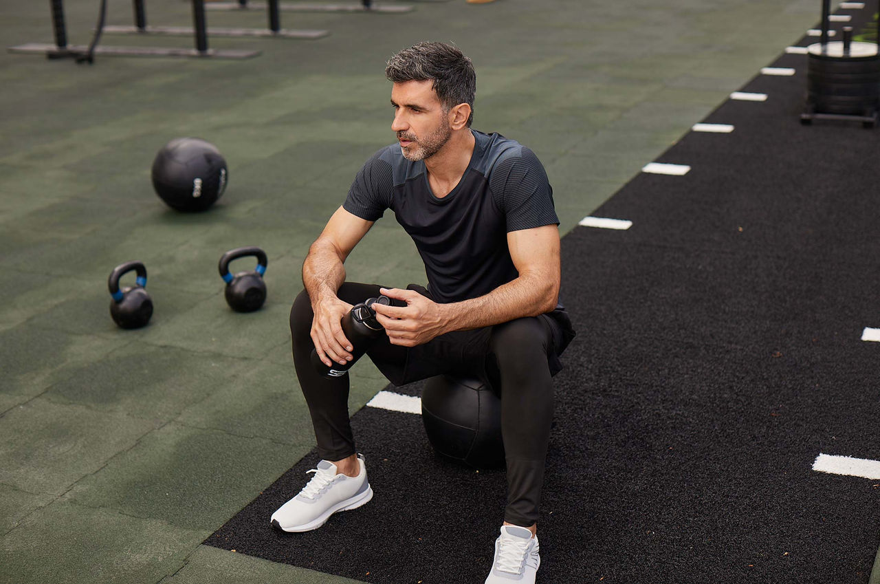A male athlete in black t-shirt, black sweatpants, and white shoes sits on medicine sits on a black medicine ball in the gym