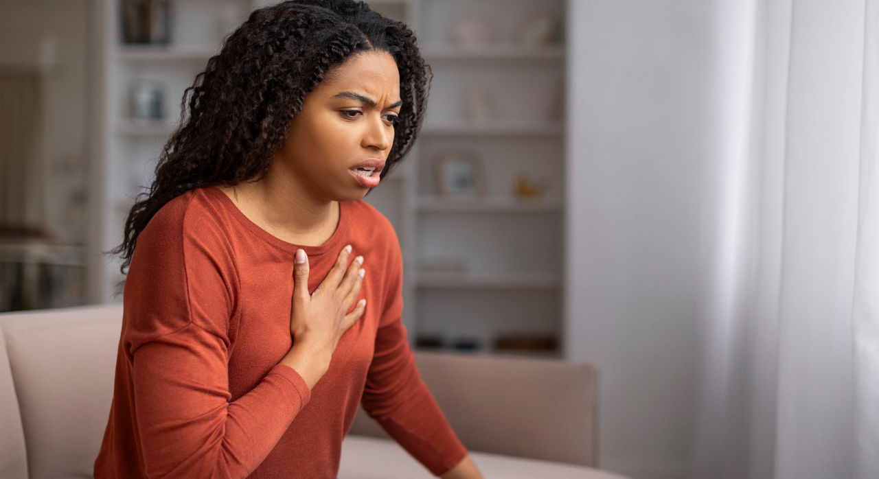 A young woman on a couch places her right hand on her chest worryingly concerned about pain