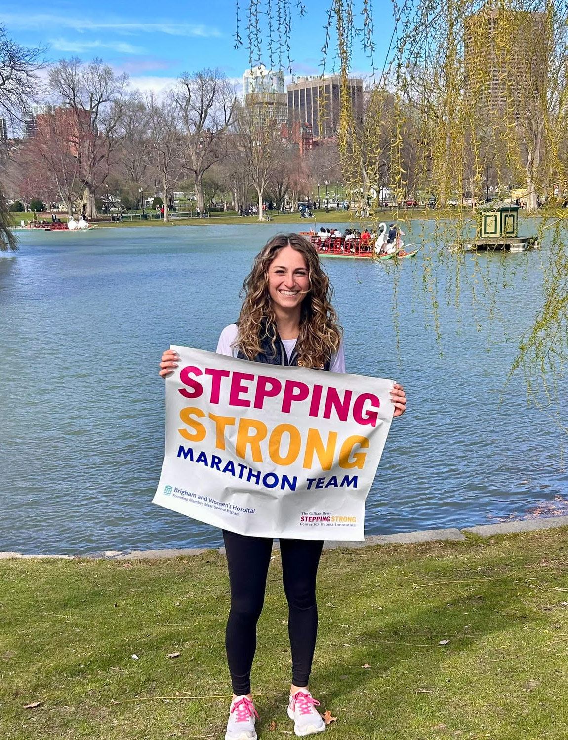 Eline Laurent holding up a poster that reads Stepping Strong Marathon Team. In the background is the pond at Boston Public Gardens with a duck boat paddling by.