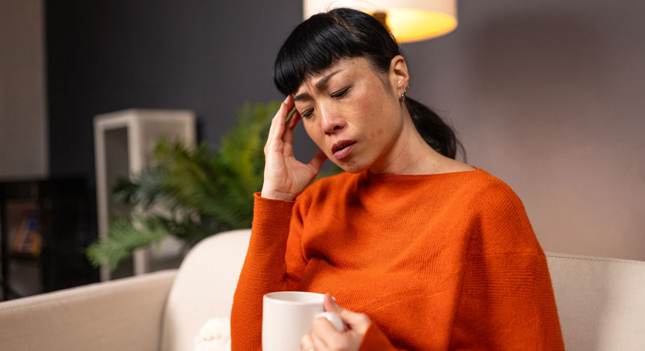 A woman with black hair sitting on her couch holding a cup of tea with her hand to her temple in pain