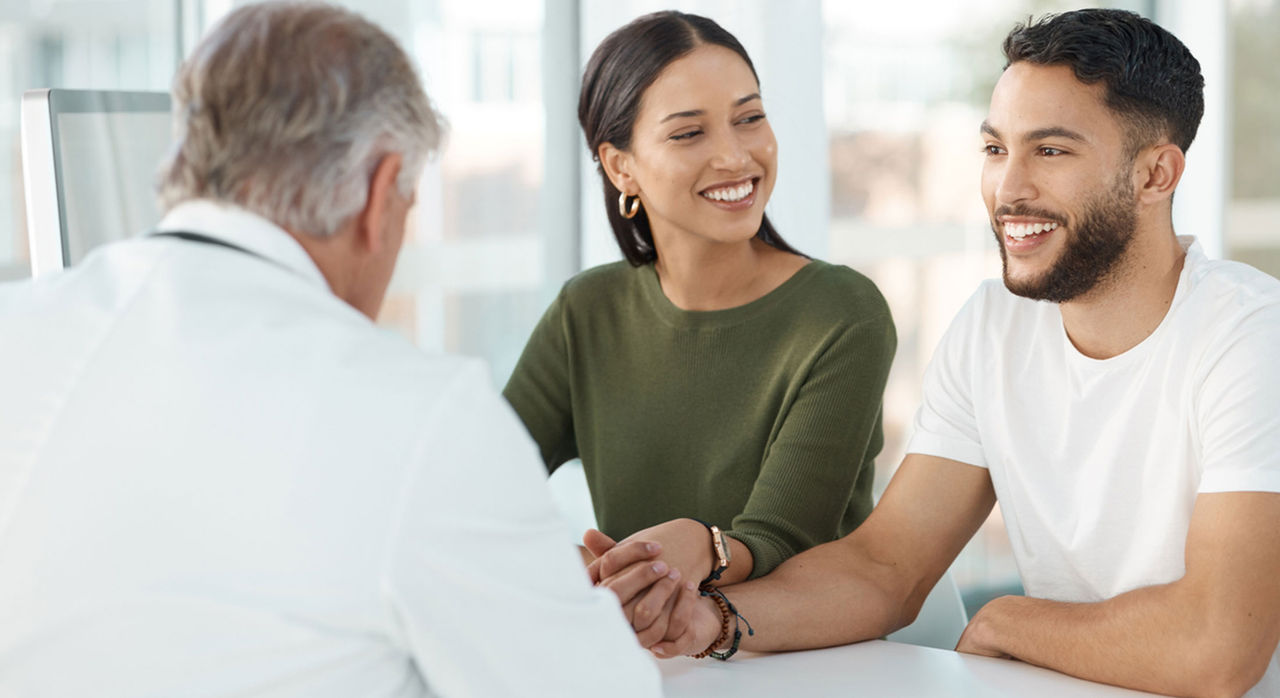 A smiling couple holds hands and speaks with an obstetrician about carrier screening.