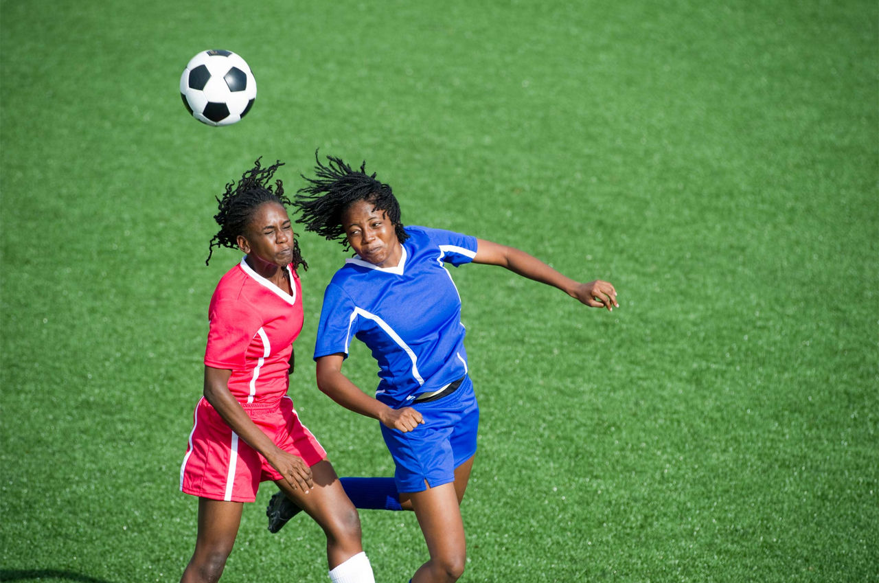 Two Black women soccer players try to head the soccer ball on the field. The player on the left is wearing a red kit, while the player on the right is wearing a blue kit.