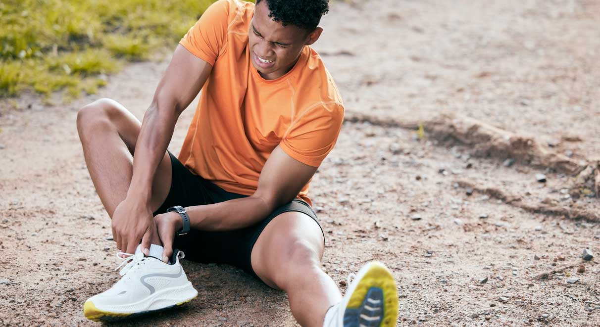 A man sits on a running path, clutching his injured ankle and grimacing.