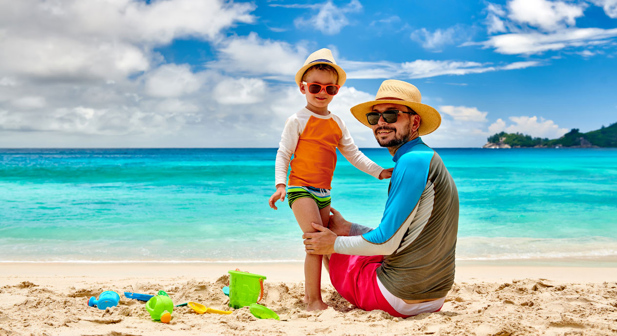 A father and his young son at the beach, wearing long -sleeved T-shirts, swimming trunks, and straw sun hats.