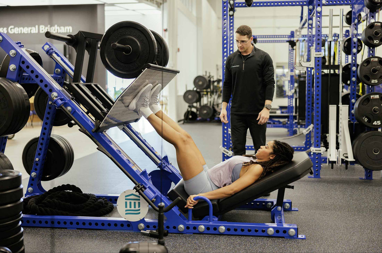 A performance coach guides an athlete using a leg press machine in a training gym