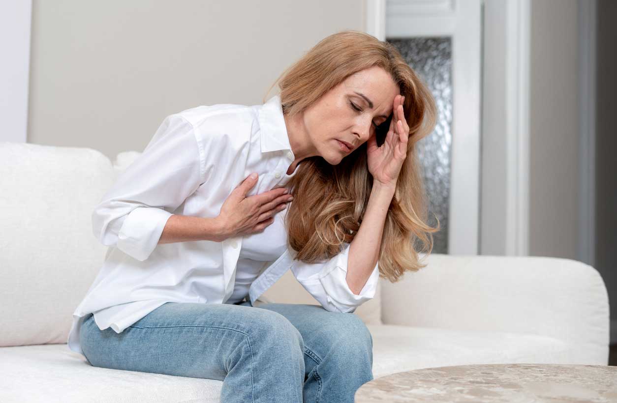 A woman sits on a couch, bent over in pain, holding one hand to her head and the other to her chest.