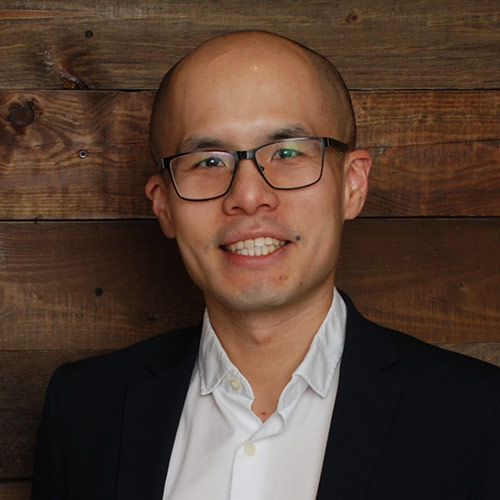 A person dressed in a white button-up shirt and dark blazer stands against a rustic wooden wall.