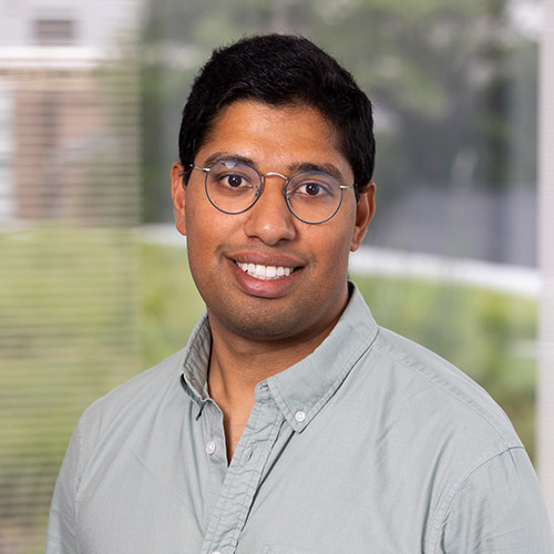 A man wearing a light gray button-down shirt is pictured indoors. The background features large windows with natural light and blurred greenery outside.