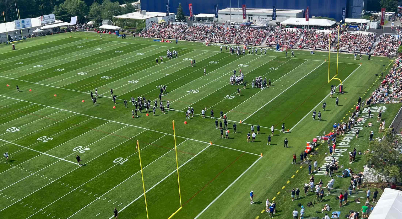 A bird's eye view of the New England Patriots field during training camp practice with players and staff on the premise and spectators looking on