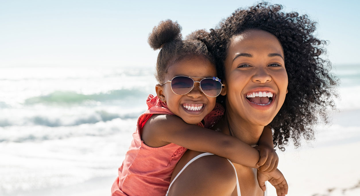 A mother and daughter romping at the beach, with the little girl riding on her mother's back.