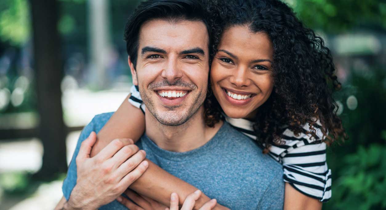 A happy couple poses for a photo outdoors, with the woman hugging the man from behind.