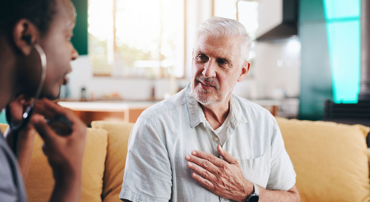 An older man touches his chest and speaks to a provider on the couch, who prepares to check his heart rate with a stethoscope.
