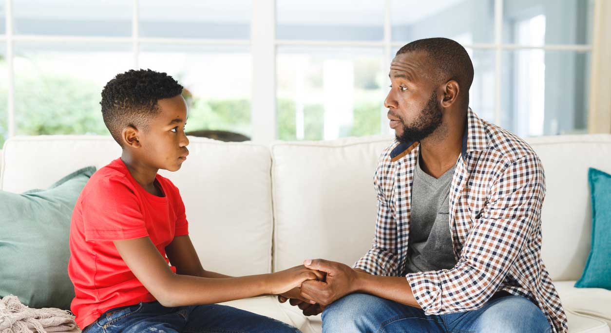 An African-American father holds his young son's hands supportively as they talk on a sofa.