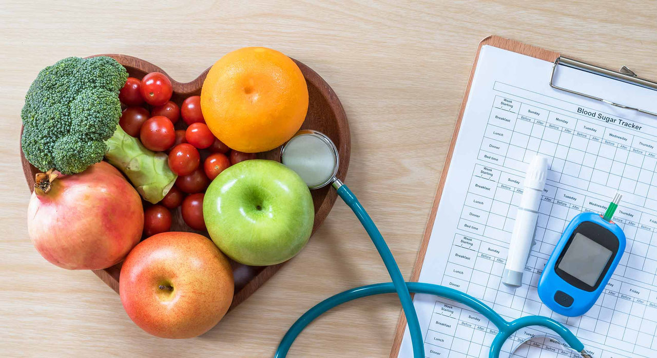 Fresh fruit and vegetables on a heart-shaped bowl next to a stethoscope, medical chart, and testing kit