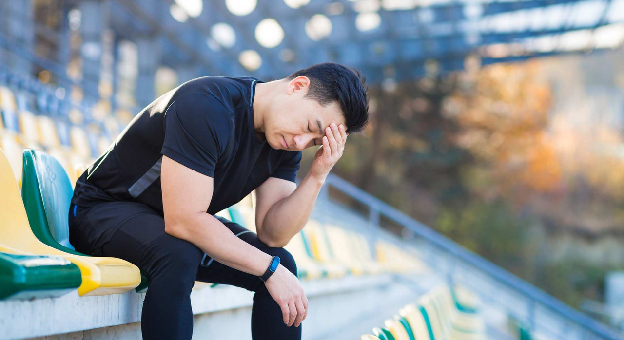 A male athlete wearing a black top and long pants sits on the bleachers while closing his eyes and resting his head on his left hand