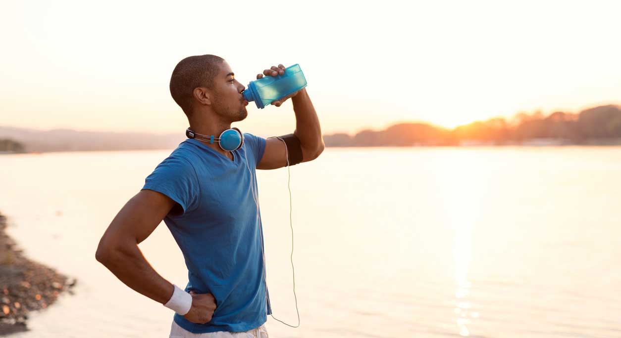 A man in workout clothes drinks from a water bottle beside a lake.