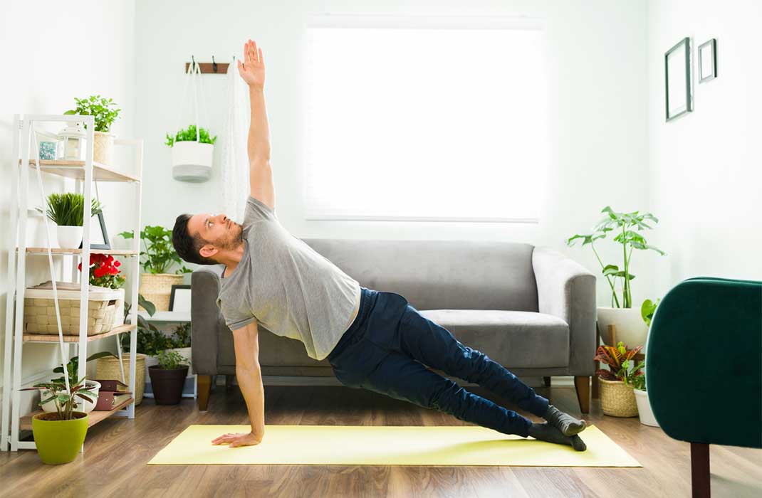 A man does a side plank in a sunny, plant-filled living room.