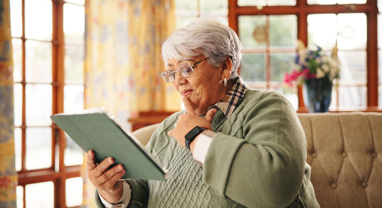 An elderly woman is seated on a comfortable sofa in a well-lit, cozy living room. She is holding a green tablet and appears engaged with the device. The room features large windows, floral curtains, and a vase of flowers in the background, creating a warm and inviting atmosphere. She is wearing a green sweater and a smartwatch on her wrist.