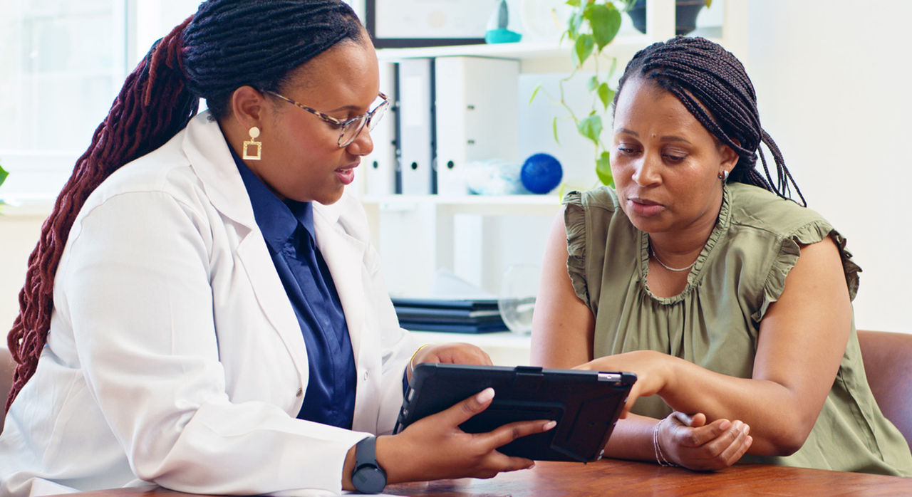 A woman of color doctor holding a tablet explaining triglycerides and cholesterol to a woman of color patient