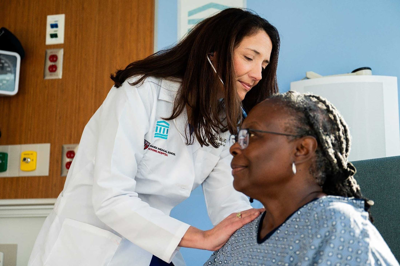 A female doctor stands and uses a stethoscope to check on a Black female patient